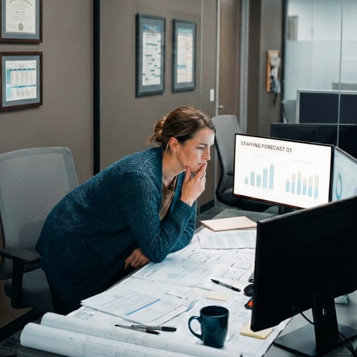 Eine Frau mittleren Alters steht nach vorn gebeugt an einem großen Schreibtisch in einem modernen Büro. Vor ihr liegen zahlreiche ausgedruckte Pläne, Tabellen und Notizen. Auf dem großen Monitor sieht man ein Diagramm mit der Überschrift „Staffing Forecast Q1“. Sie stützt das Kinn auf die Hand und wirkt konzentriert, während sie Personal- und Kapazitätsdaten analysiert. Die Szene zeigt den Arbeitsalltag einer Logistikmanagerin, die Entscheidungen für das Fahrer-Recruiting trifft und Wege sucht, schneller passende LKW-Fahrer zu finden – etwa durch effizientere Job-Matching-Lösungen.